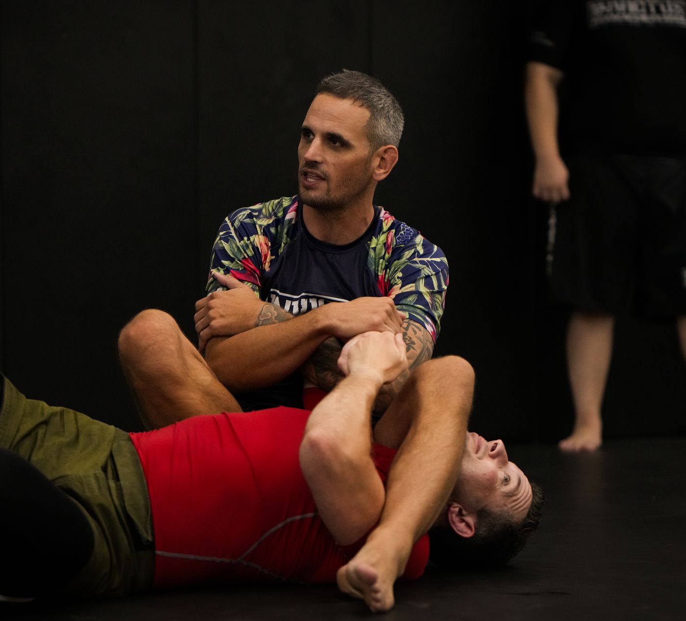 Coach Nate sits on a student's chest, arms folded, talking to the class.