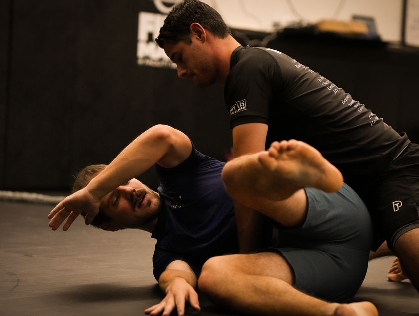 A coach in a black rash guard works a leg position on an adult student on the mats.
